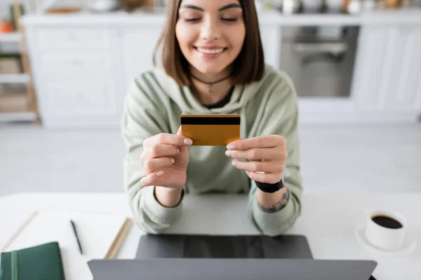 Femme souriante tenant une carte de crédit près d'un ordinateur portable et café sur la table à la maison — Photo de stock