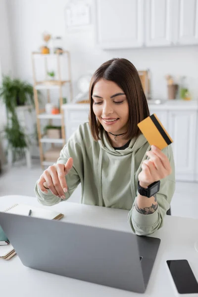 Положительная женщина держит кредитную карту во время покупок в Интернете на ноутбуке дома — стоковое фото