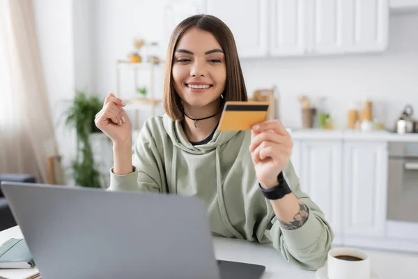 Mujer tatuada alegre mirando la tarjeta de crédito cerca del ordenador portátil y el café en la cocina - foto de stock