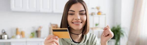 Усміхнена брюнетка, що тримає розмиту кредитну картку вдома, банер — стокове фото