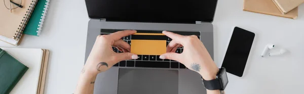Vue du dessus de la femme tatouée tenant la carte de crédit près des gadgets et des ordinateurs portables sur la table, bannière — Photo de stock