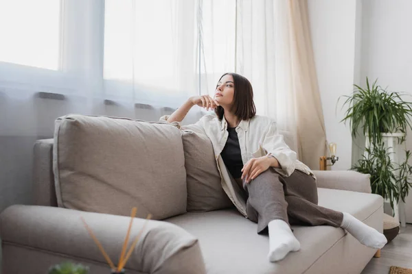Молода жінка в повсякденному одязі дивиться далеко, сидячи на дивані вдома — стокове фото