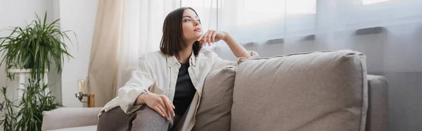 Bruna donna in abiti casual guardando lontano mentre seduto sul divano a casa, banner — Foto stock
