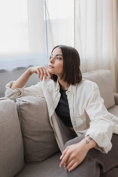 Jovem tatuado mulher olhando para longe enquanto sentado no sofá em casa — Fotografia de Stock