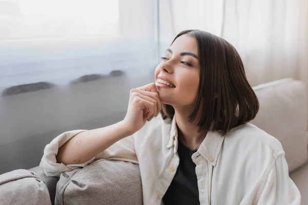 Fröhliche junge Frau im Hemd schaut weg, während sie zu Hause auf der Couch sitzt — Stockfoto