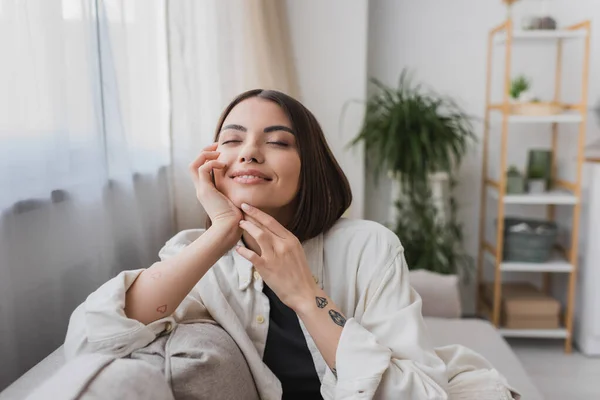 Щаслива татуйована жінка торкається обличчя, сидячи на дивані вдома — стокове фото