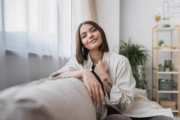 Pleased young woman looking at camera while sitting on blurred couch at home — Stock Photo