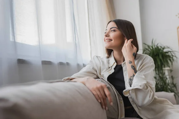 Позитивна молода жінка торкається волосся, сидячи на дивані вдома — стокове фото