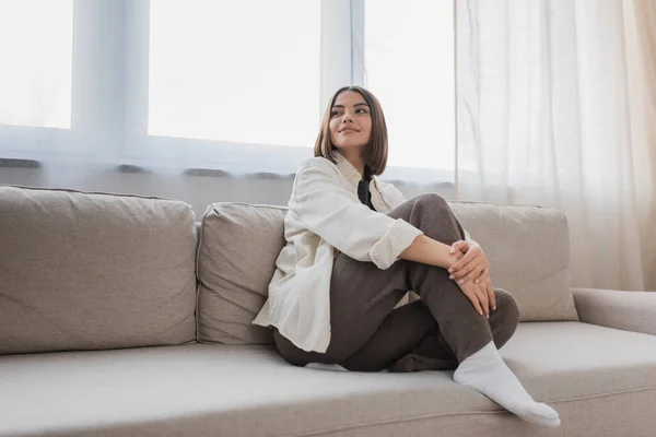 Lächelnde junge Frau schaut weg, während sie zu Hause auf der Couch sitzt — Stockfoto