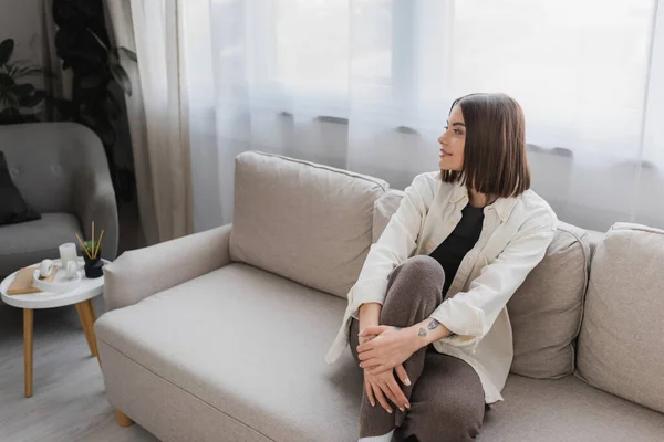 Giovane donna bruna sorridente mentre siede sul divano in soggiorno — Foto stock