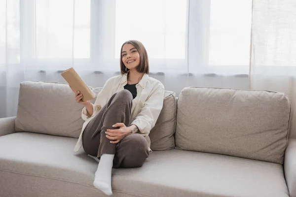 Весела молода жінка тримає книгу, сидячи на дивані у вітальні — стокове фото