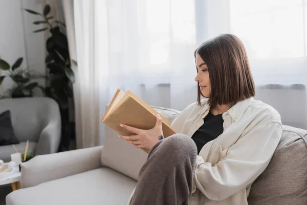 Vue latérale de la femme brune tenant le livre assis sur le canapé à la maison — Photo de stock