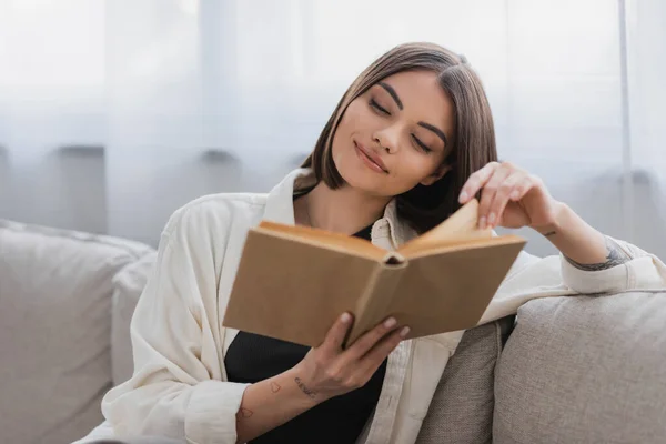 Радостная татуированная женщина читает размытую книгу в гостиной — стоковое фото