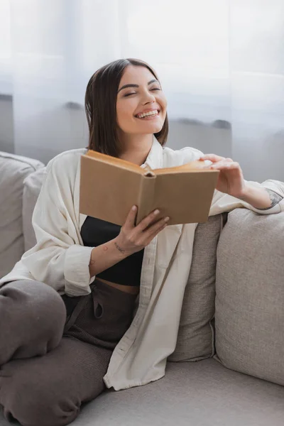 Fröhliche junge Frau hält Buch in der Hand, während sie auf Couch im Wohnzimmer sitzt — Stockfoto