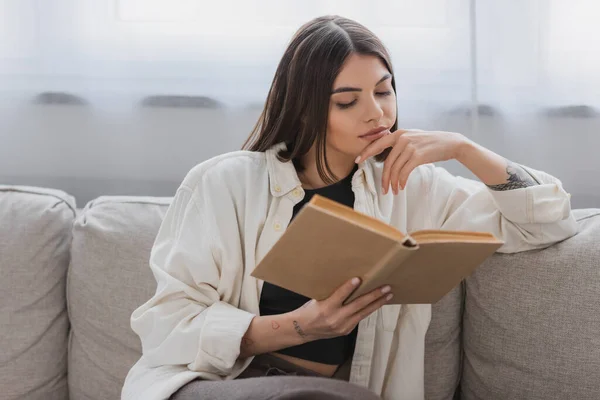 Mujer joven tatuada leyendo libro en el sofá en la sala de estar - foto de stock