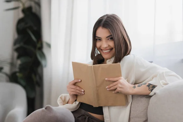Positiv tätowierte Frau hält Buch in der Hand und blickt in die Kamera im Wohnzimmer — Stockfoto