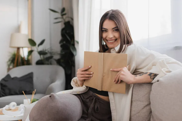 Femme brune souriante tenant le livre et regardant la caméra sur le canapé à la maison — Photo de stock