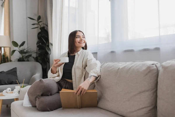 Femme positive tenant tasse de café et réserver tout en se reposant sur le canapé à la maison — Photo de stock