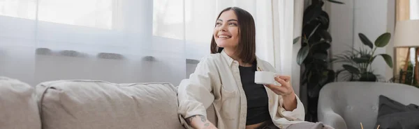 Overjoyed woman holding cup of coffee while sitting on couch at home, banner — Stock Photo