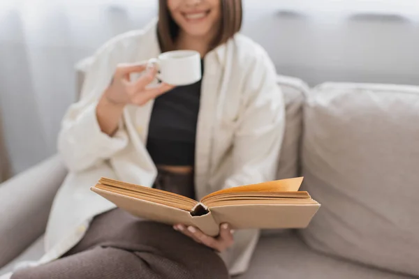Cropped view of blurred woman holding coffee cup and reading book on couch at home — Stock Photo
