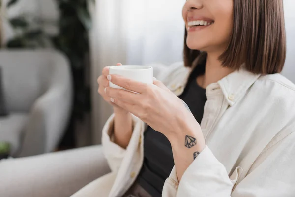 Vista cortada de mulher tatuada despreocupada segurando xícara de café na sala de estar — Fotografia de Stock