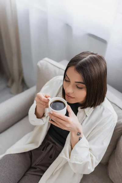 Вид сверху на татуированную женщину, держащую чашку кофе, сидя на диване — стоковое фото