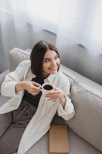 Vista de ángulo alto de mujer morena alegre sosteniendo taza de café cerca de libro en sofá en sala de estar - foto de stock