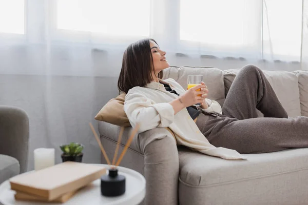 Vista lateral da mulher satisfeita segurando vidro de suco de laranja no sofá em casa — Fotografia de Stock