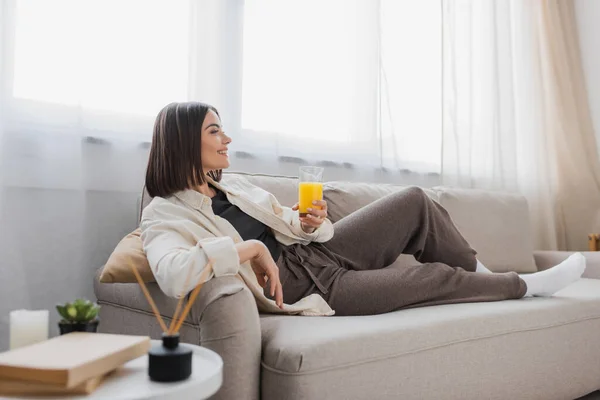 Vista lateral de una mujer morena sonriente sosteniendo un vaso de jugo de naranja mientras está sentada en el sofá en casa - foto de stock