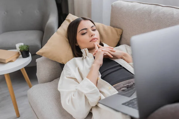 Focused brunette freelancer looking at laptop while lying on couch at home — Stock Photo