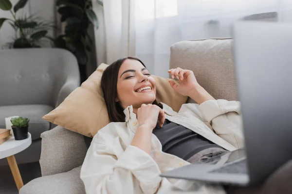 Cheerful brunette woman with blurred laptop while lying on couch at home — Stock Photo