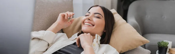Jeune femme positive couchée sur le canapé près d'un ordinateur portable flou à la maison, bannière — Photo de stock