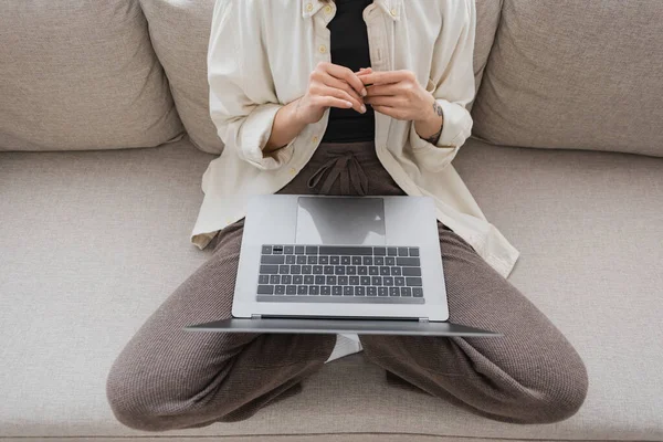 Обрезанный вид татуированной женщины в повседневной одежде, держащей ноутбук, сидя дома на диване — стоковое фото