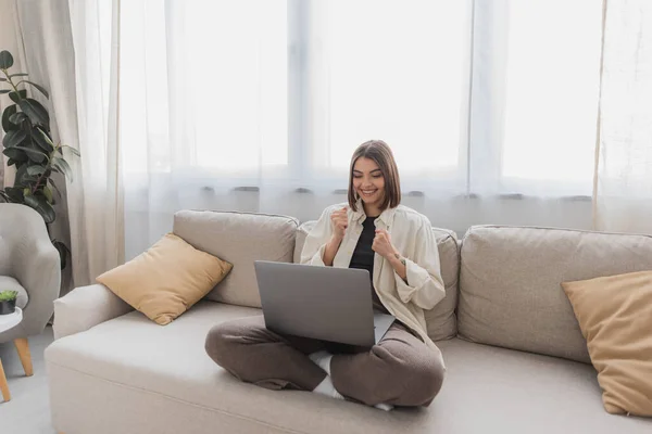 Aufgeregte Freiberuflerin zeigt Ja-Geste und schaut auf Laptop zu Hause auf Couch — Stockfoto