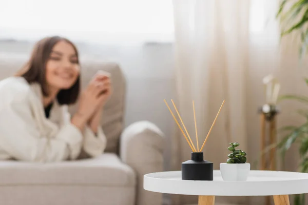 Planta y bambú palos perfumados en la mesa de café cerca de la mujer borrosa en el sofá en casa - foto de stock