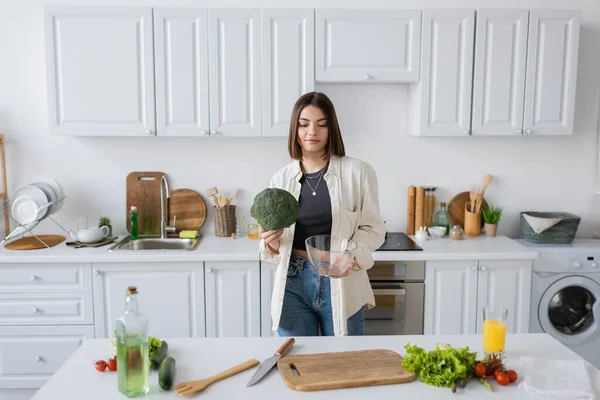 Brunette woman holding broccoli and bowl near vegetables in kitchen — Stock Photo