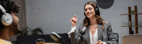 Sorrindo e encantador anfitrião de rádio morena em fones de ouvido e blazer cinza gesticulando enquanto conversa com o homem indiano borrado perto de microfones profissionais e café para ir em estúdio de radiodifusão, banner — Fotografia de Stock