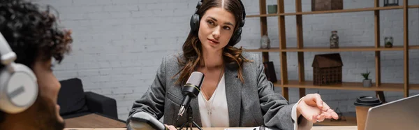 Charming radio host in headphones and grey blazer pointing at laptop near coffee to go and professional microphone during interview with blurred indian guest in broadcasting studio, banner — Stock Photo