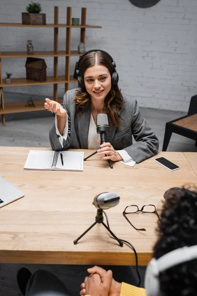 Smiling and charming announcer in headphones talking and gesturing near microphone, smartphone and notebook with pen while recording interview with blurred indian man in radio studio — Stock Photo