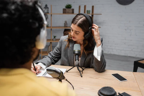 Charming broadcaster in headphones and grey blazer writing in notebook near microphones, smartphone with blank screen and paper cup during interview with blurred indian man in radio studio — Stock Photo