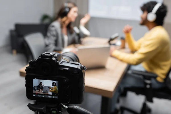 Selective focus of professional digital camera recording how interviewer talking to young indian guest while sitting together near laptop and microphones on blurred background in studio — Stock Photo