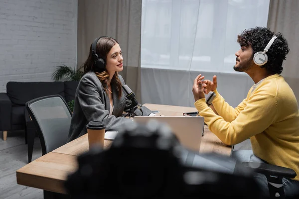 Side view of smiling brunette radio host in headphones and blazer looking at curly indian man in yellow jumper talking and gesturing during interview near laptop and paper cup in modern radio studio — Stock Photo