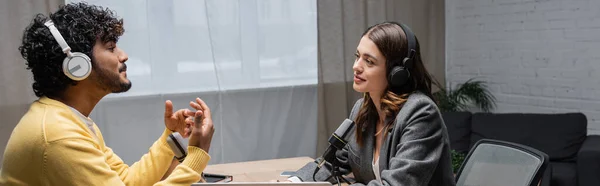 Side view of young and curly indian man in headphones and yellow jumper talking and gesturing near microphones and smiling brunette interviewer wearing grey blazer in radio studio, banner — Stock Photo