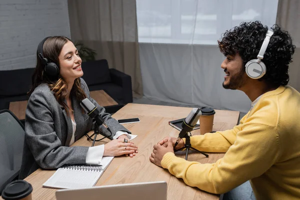 Happy and charming radio host in headphones and young curly indian man in yellow jumper talking near microphones, paper cups, smartphone and notebooks in professional studio — Stock Photo
