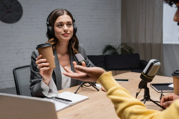 Brunette interviewer in headphones holding paper cup near professional microphones, notebooks and smartphone while looking at indian guest in yellow jumper gesturing on blurred foreground in studio — Stock Photo