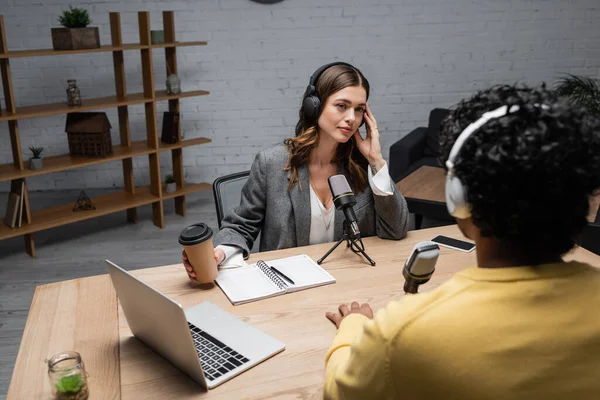 Tätowierte brünette Interviewerin mit Kopfhörer, Imbissgetränk in der Hand und Blick auf verschwommenen indischen Mann in der Nähe von Laptop, Mikrofonen, Notebook und Handy auf dem Tisch im Sendestudio — Stockfoto