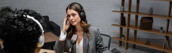 Brunette radio host in grey blazer adjusting headphones during interview with blurred indian man near microphone and paper cup with wooden rack on background in broadcasting studio, banner — Stock Photo