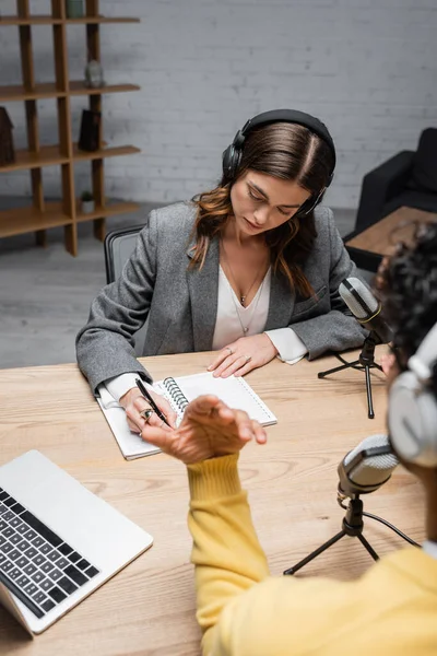 Brunette broadcaster in headphones and grey blazer writing in notebook near microphones and laptop while indian man gesturing on blurred foreground during interview in radio studio — Stock Photo