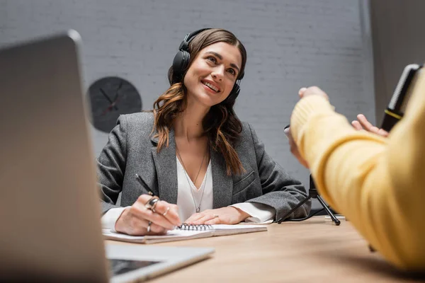 Happy brunette podcaster in headphones and grey blazer writing in notebook near microphones and looking at blurred man gesturing near laptop during interview in broadcasting studio — Stock Photo