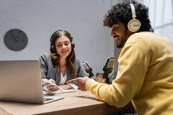 Charming radio host in grey blazer showing smartphone to young indian man in headphones and yellow jumper pointing at laptop near professional microphones in broadcasting studio — Stock Photo